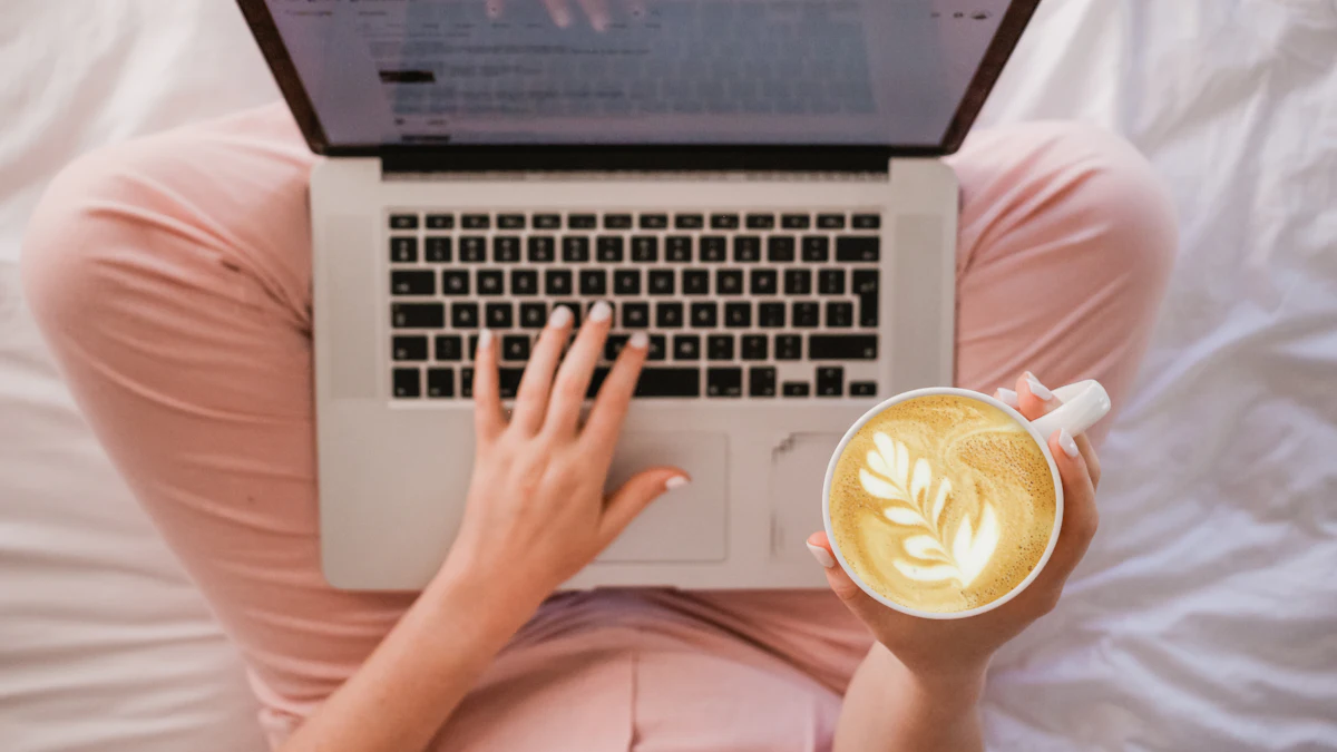 A person working on a laptop in bed with a cup of coffee in one hand