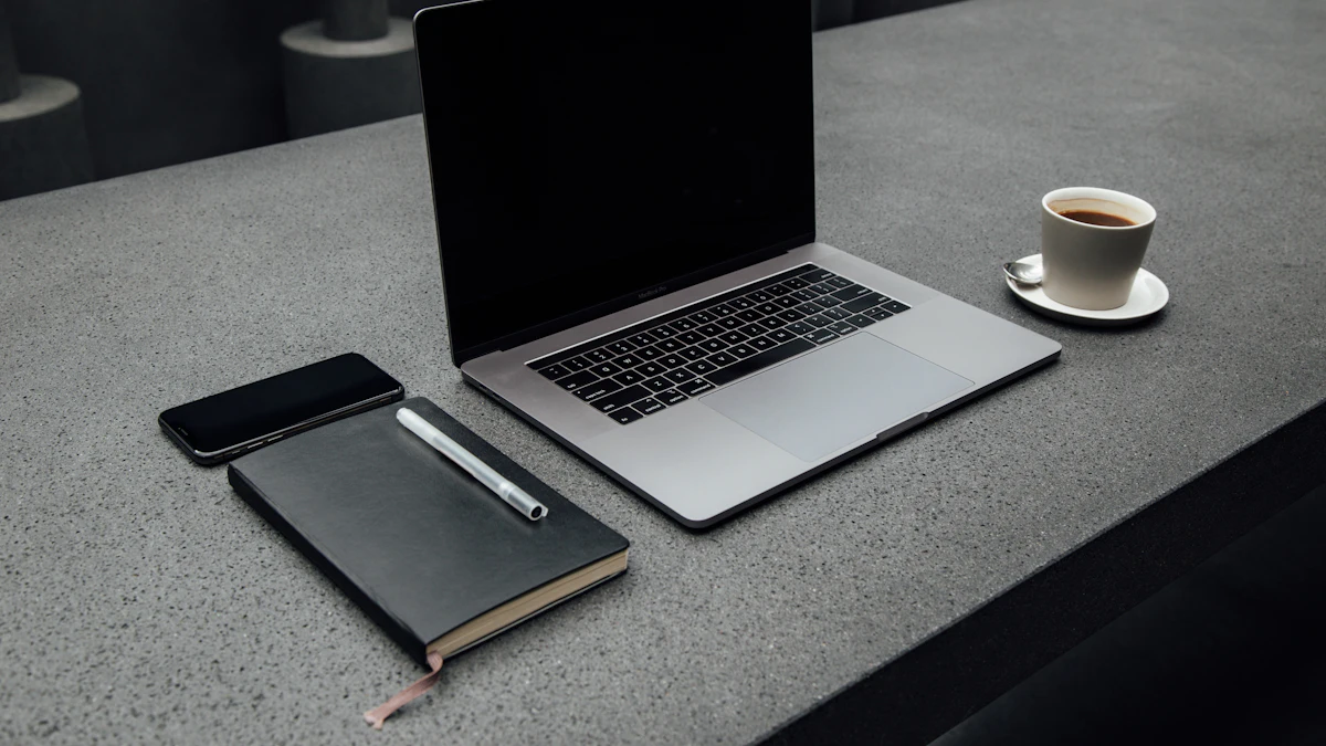 A laptop, phone, notebook and pen, and a cup of coffee on a grey desk