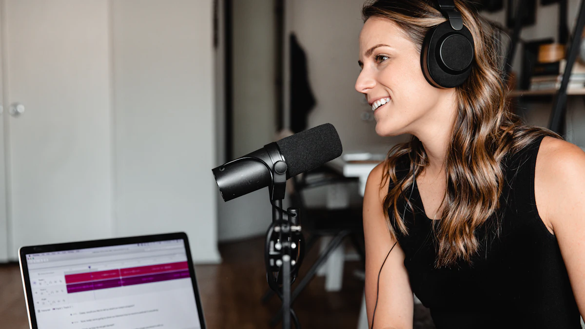 A woman in headphones smiling beside a microphone on a stand, with a laptop open in front of her