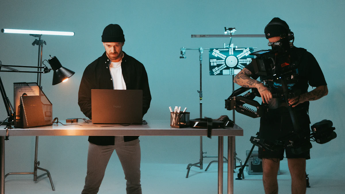 A man working at a laptop on a desk in a blue-lit studio while a camera operator films beside him