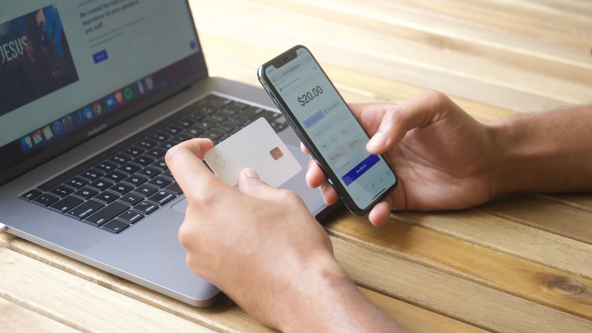 Hands holding a bank card and a phone showing a payment screen, beside an open laptop